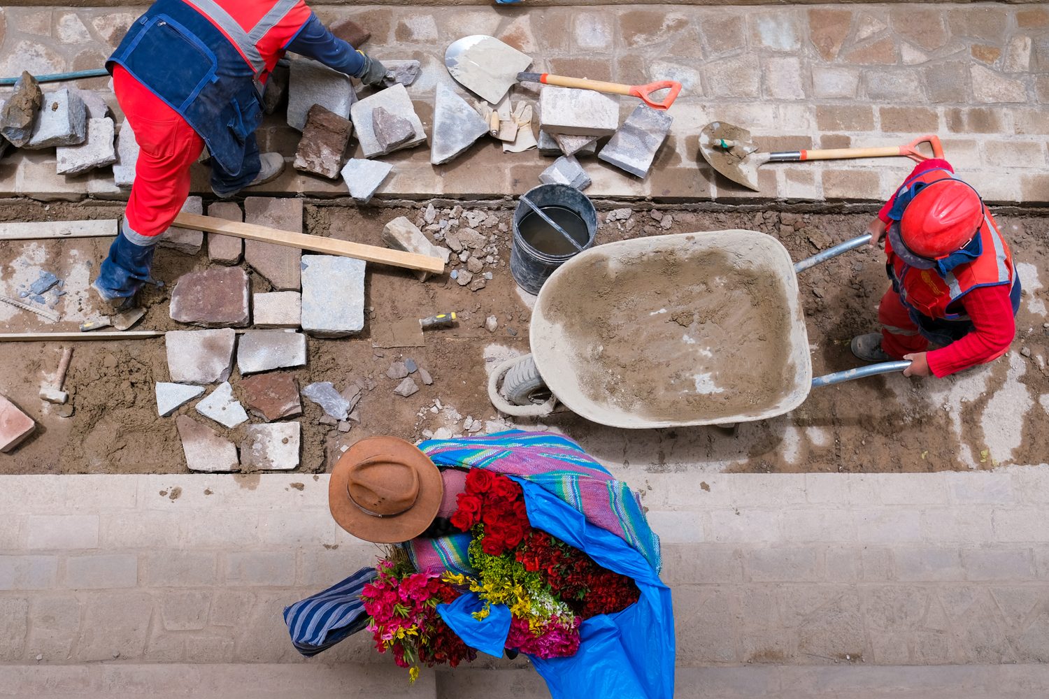 Overhead view of workers laying stone, a flower-seller passing below