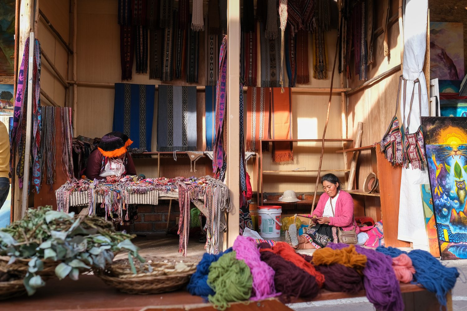 Two weavers at work in a market stall hung with textiles