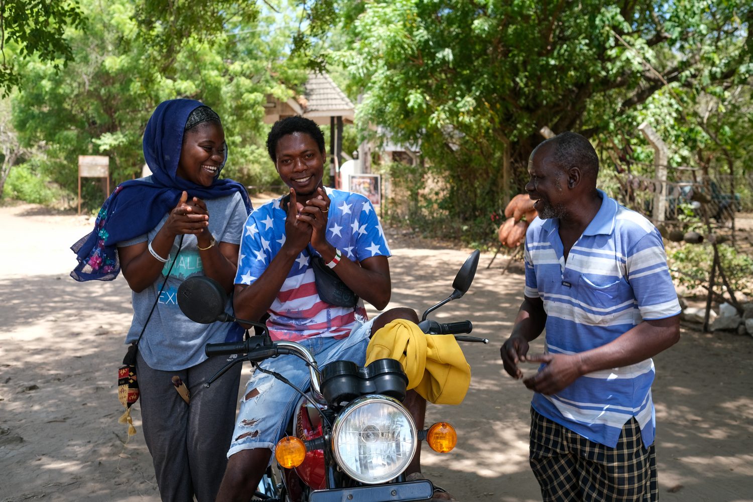 Three people laughing together around a motorbike on a dirt road