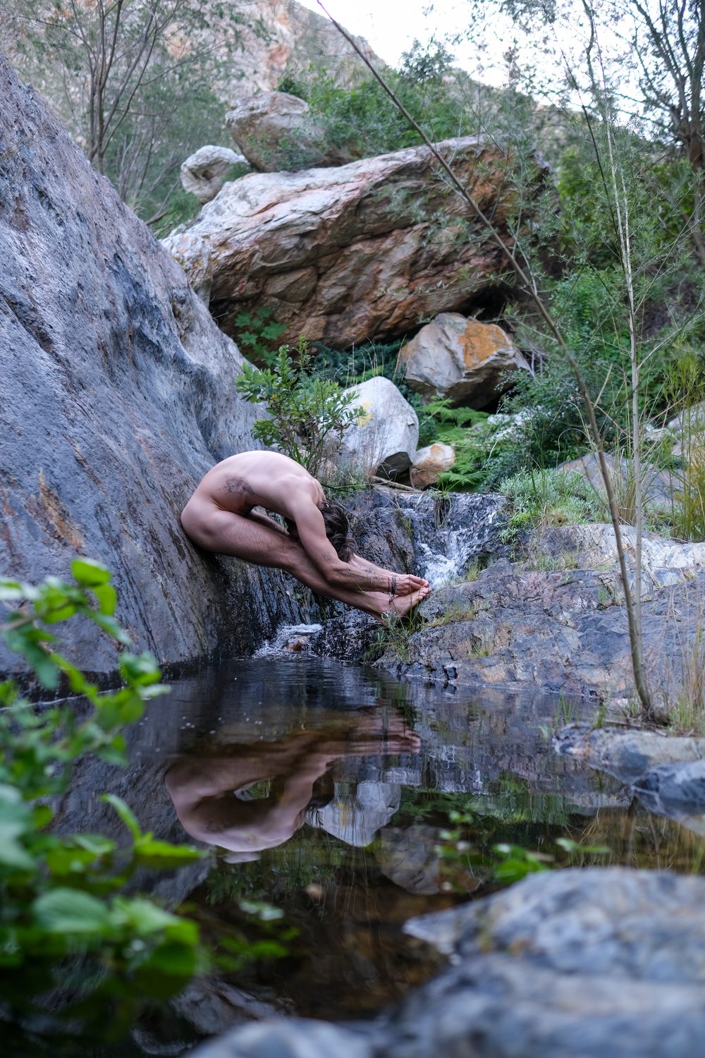 A figure folded forward beside a mountain rock pool, mirrored in still water